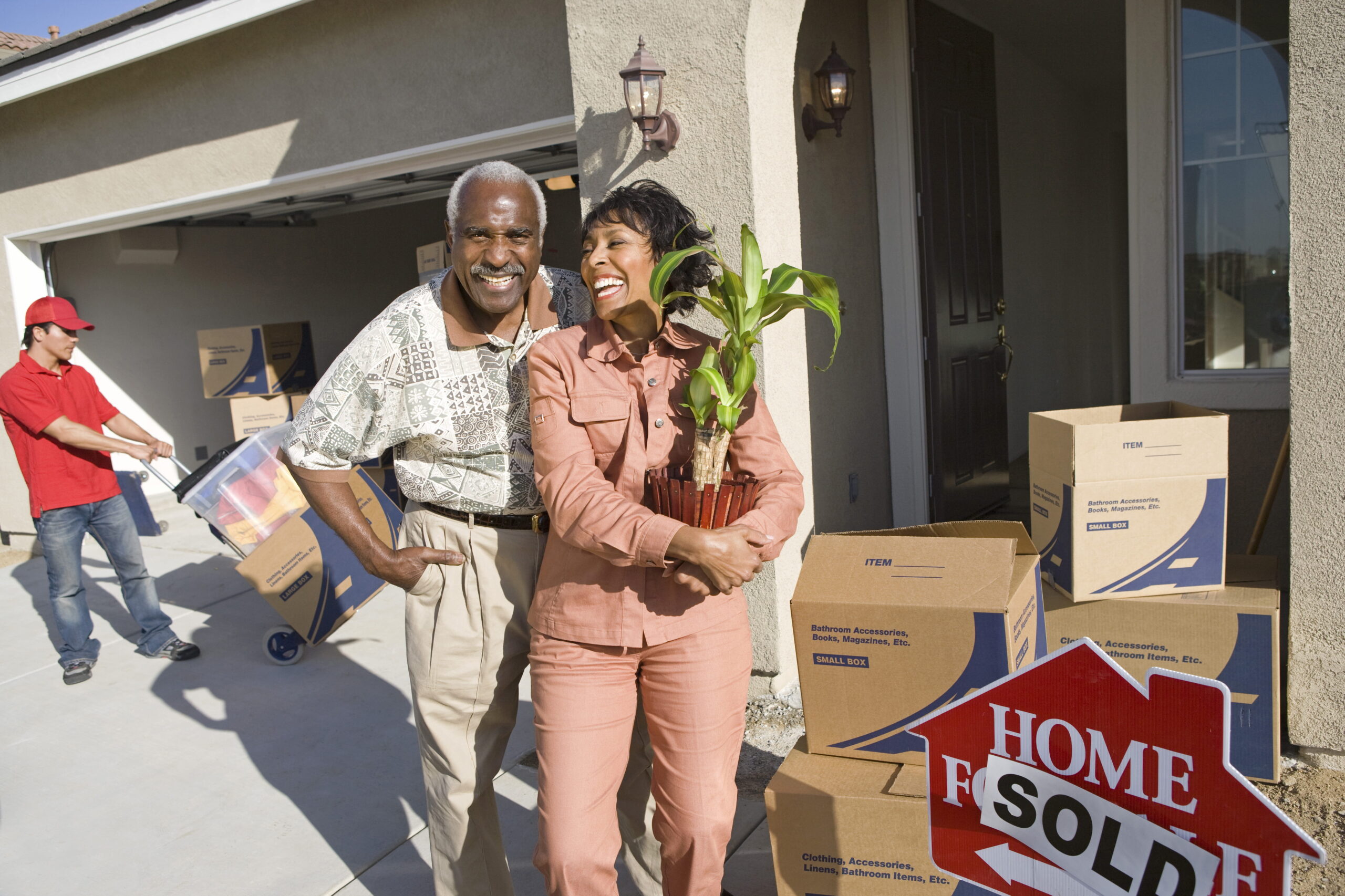 Elderly couple happily stand outside their new home, holding a plant. Moving boxes and a "Home Sold" sign are visible; a worker carries more boxes.