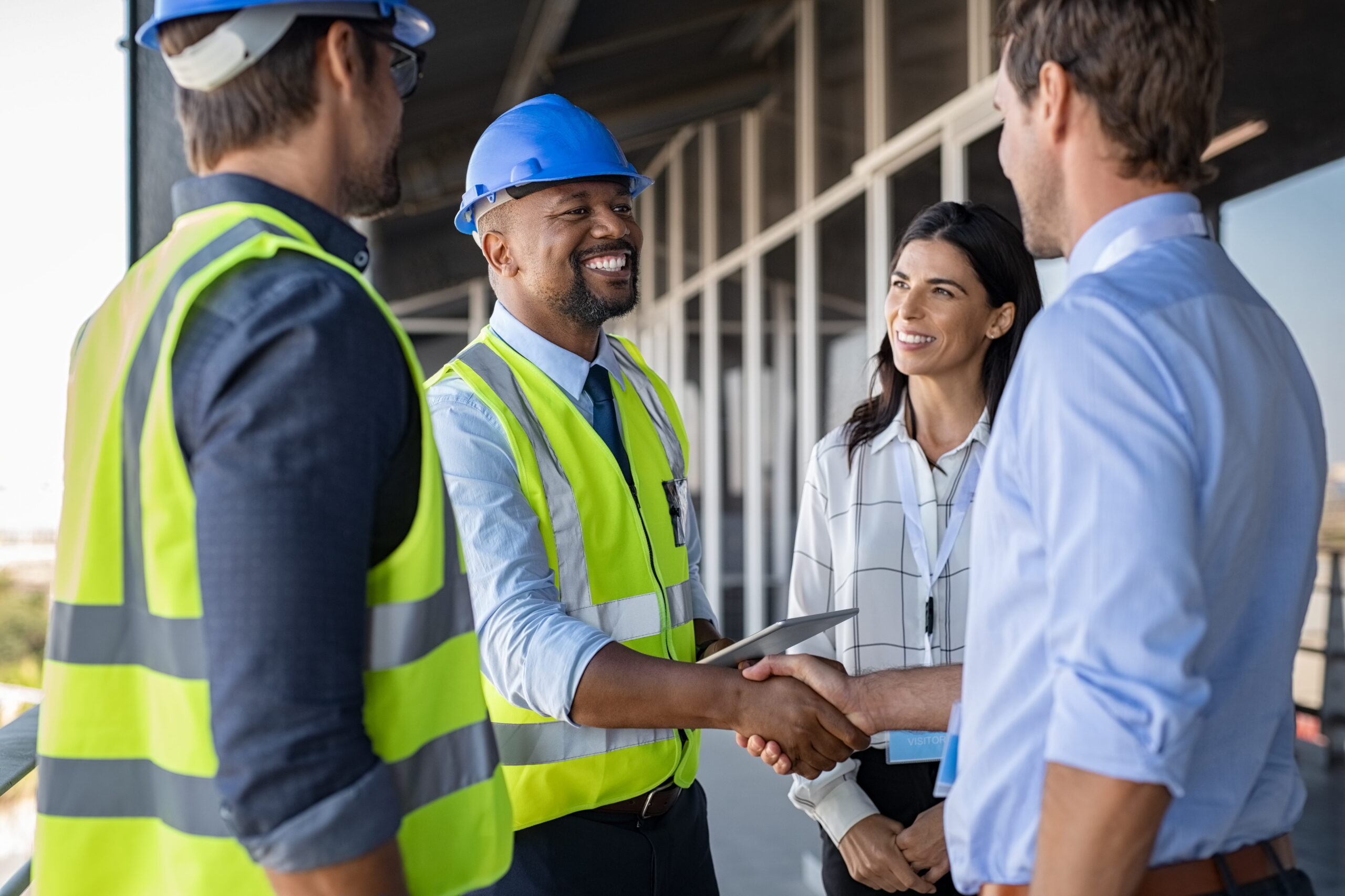 A construction worker in a safety vest and hard hat shakes hands with a smiling professional in a shirt. Two colleagues observe, conveying teamwork.